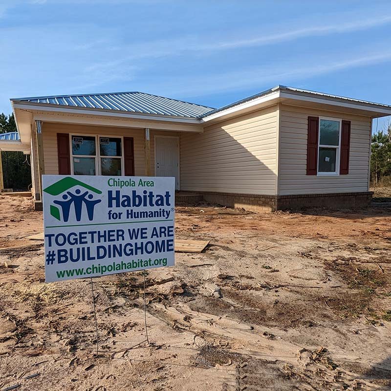 a home under construction sits on a dirt lot behind a sign that reads Chipola Area Habitat for Humanity together we are #buildinghome